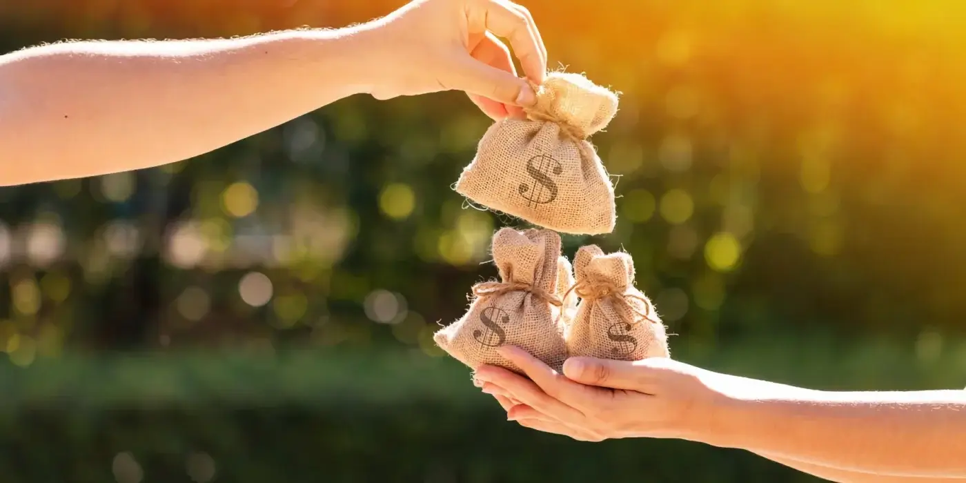 Two hands pass small burlap money bags with dollar signs between them outdoors, with sunlight and greenery in the background.