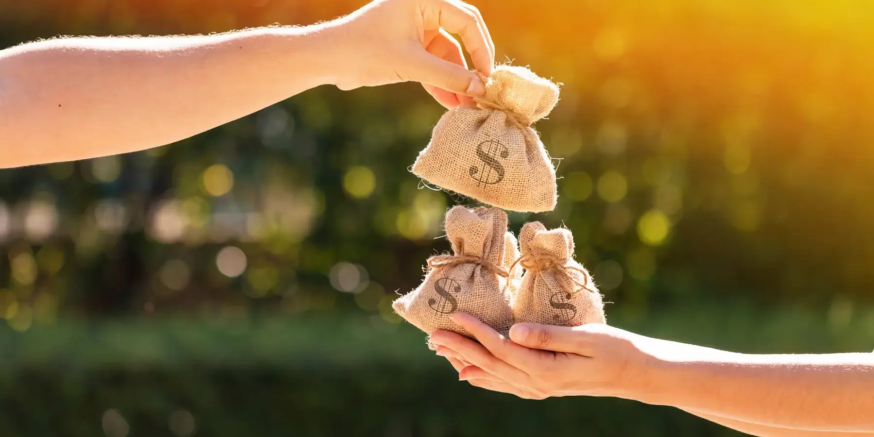 Two hands pass small burlap money bags with dollar signs between them outdoors, with sunlight and greenery in the background.