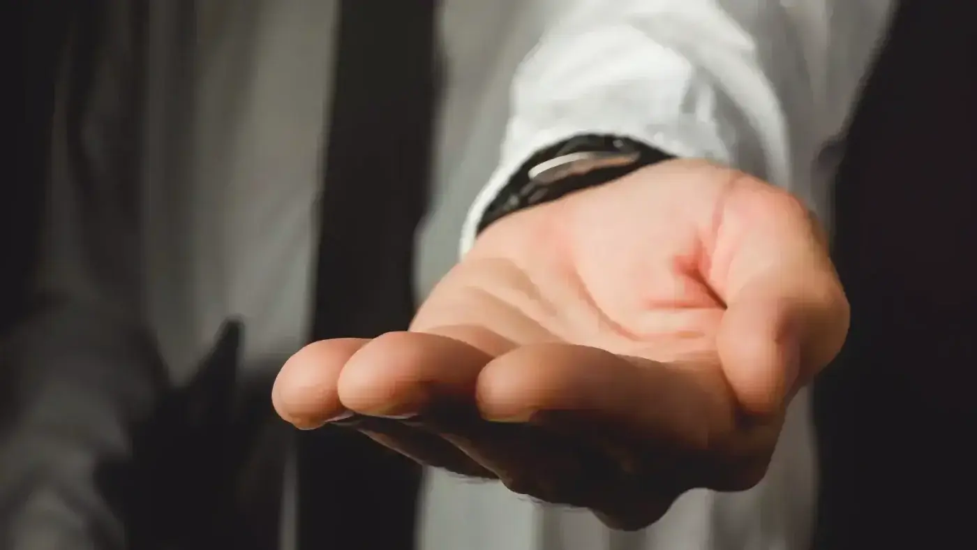 A close-up of a person’s open hand extended forward, palm up, wearing a white shirt and a black watch, suggesting a gesture of asking or offering.