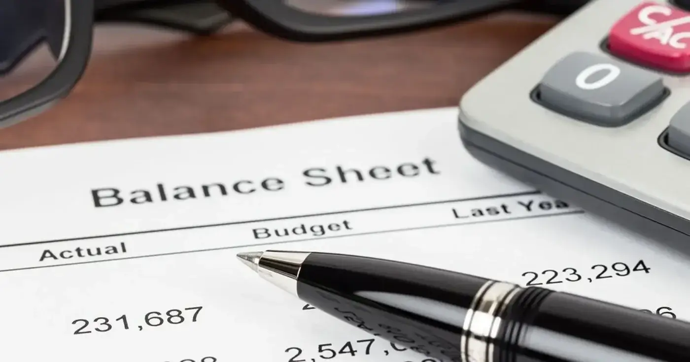 Close-up of a balance sheet financial statement with rows of numbers, a pen resting on top, and part of a calculator and a pair of glasses visible in the background.
