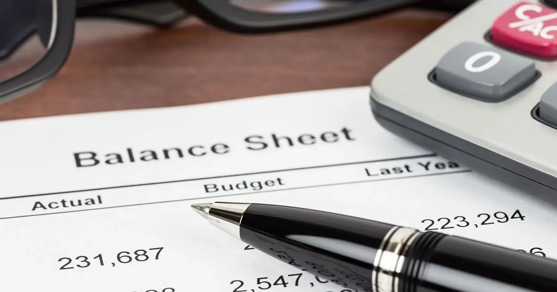 Close-up of a balance sheet financial statement with rows of numbers, a pen resting on top, and part of a calculator and a pair of glasses visible in the background.