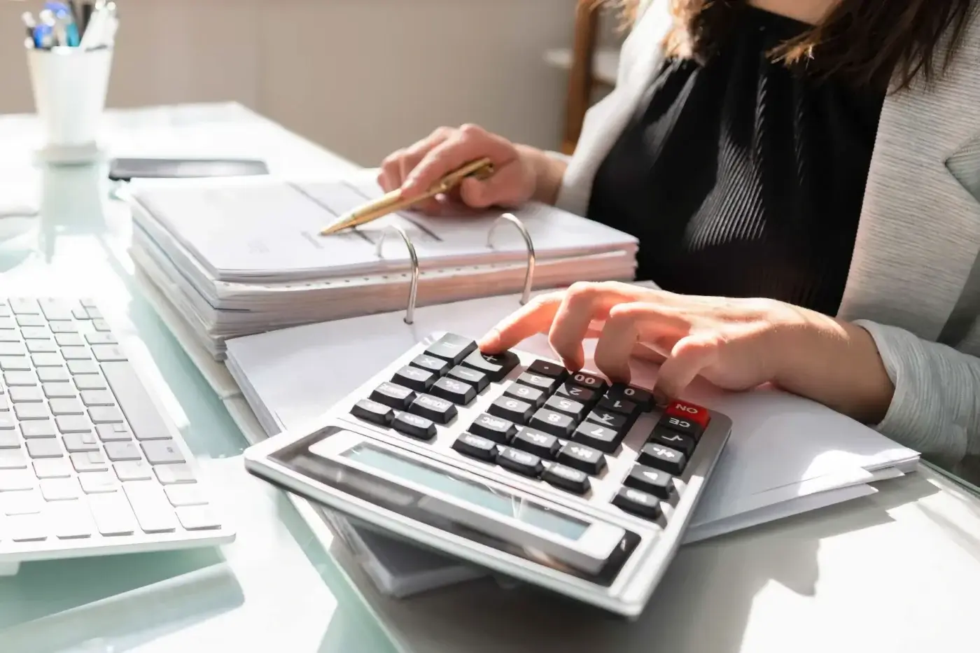 A person in business attire uses a calculator and holds a pen over a stack of documents on a desk, next to a computer keyboard.