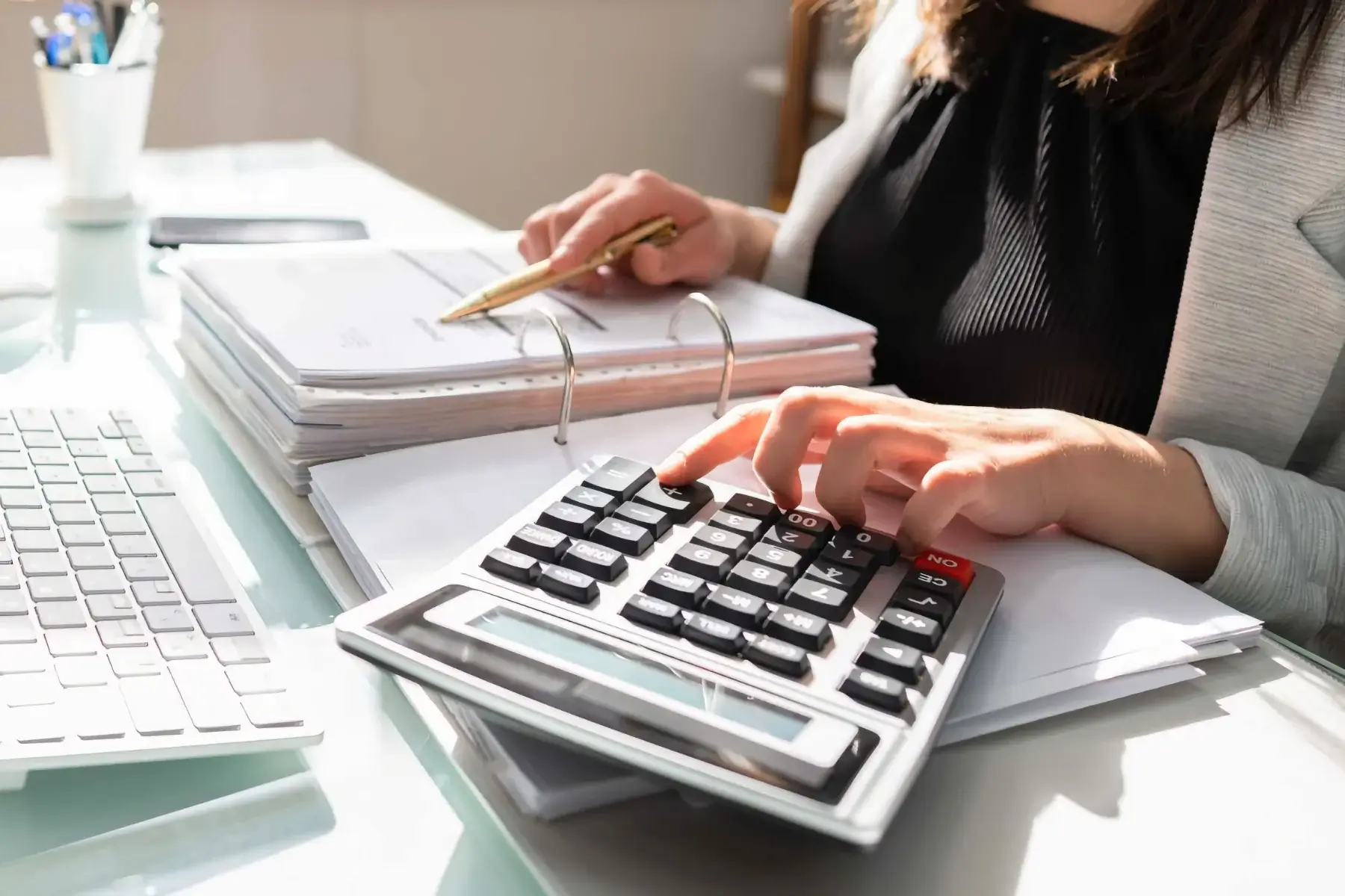 A person in business attire uses a calculator and holds a pen over a stack of documents on a desk, next to a computer keyboard.