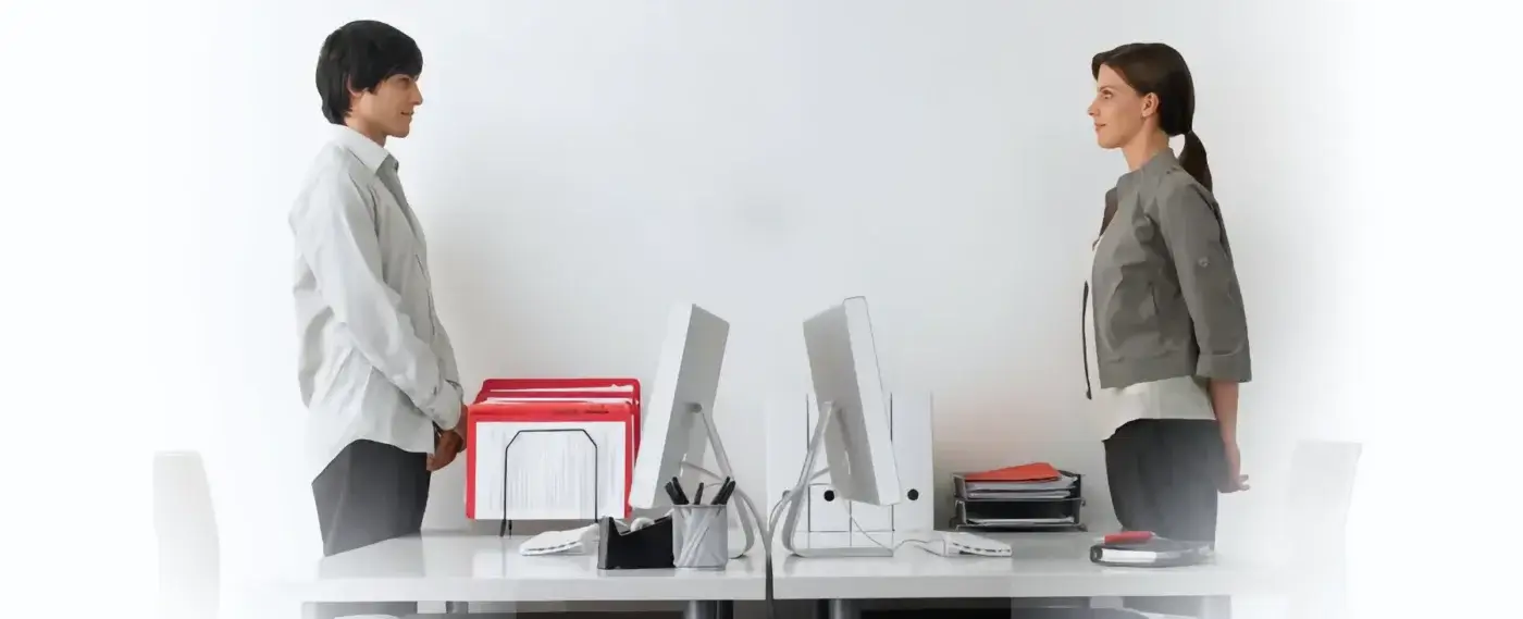 Two people stand facing each other across a desk with computers, office supplies, and file folders in a modern, minimal office—perfect for a small business accountant or bookkeeper.