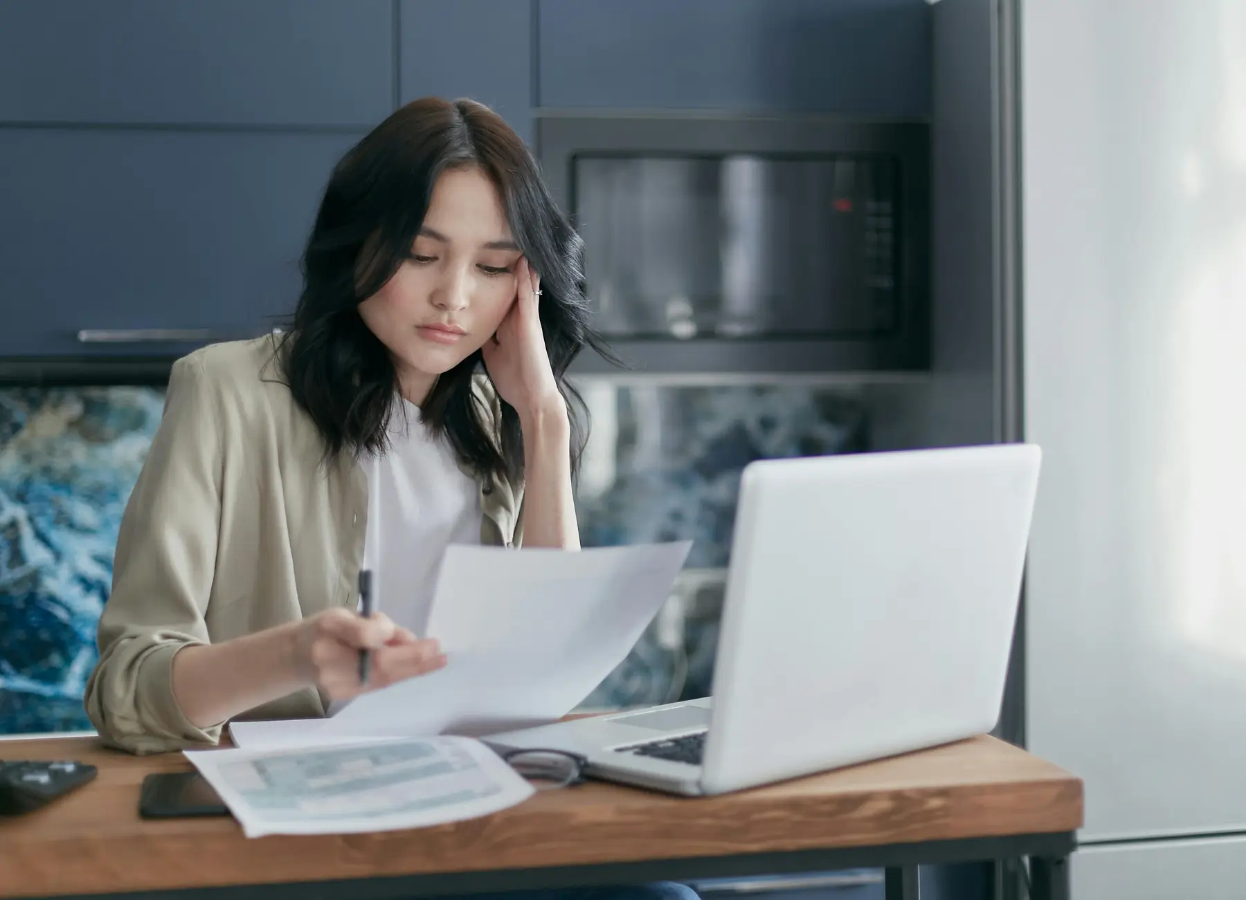 A woman sits at a table, holding documents and using a laptop. She appears focused, with a pen in hand. A microwave is visible in the background.