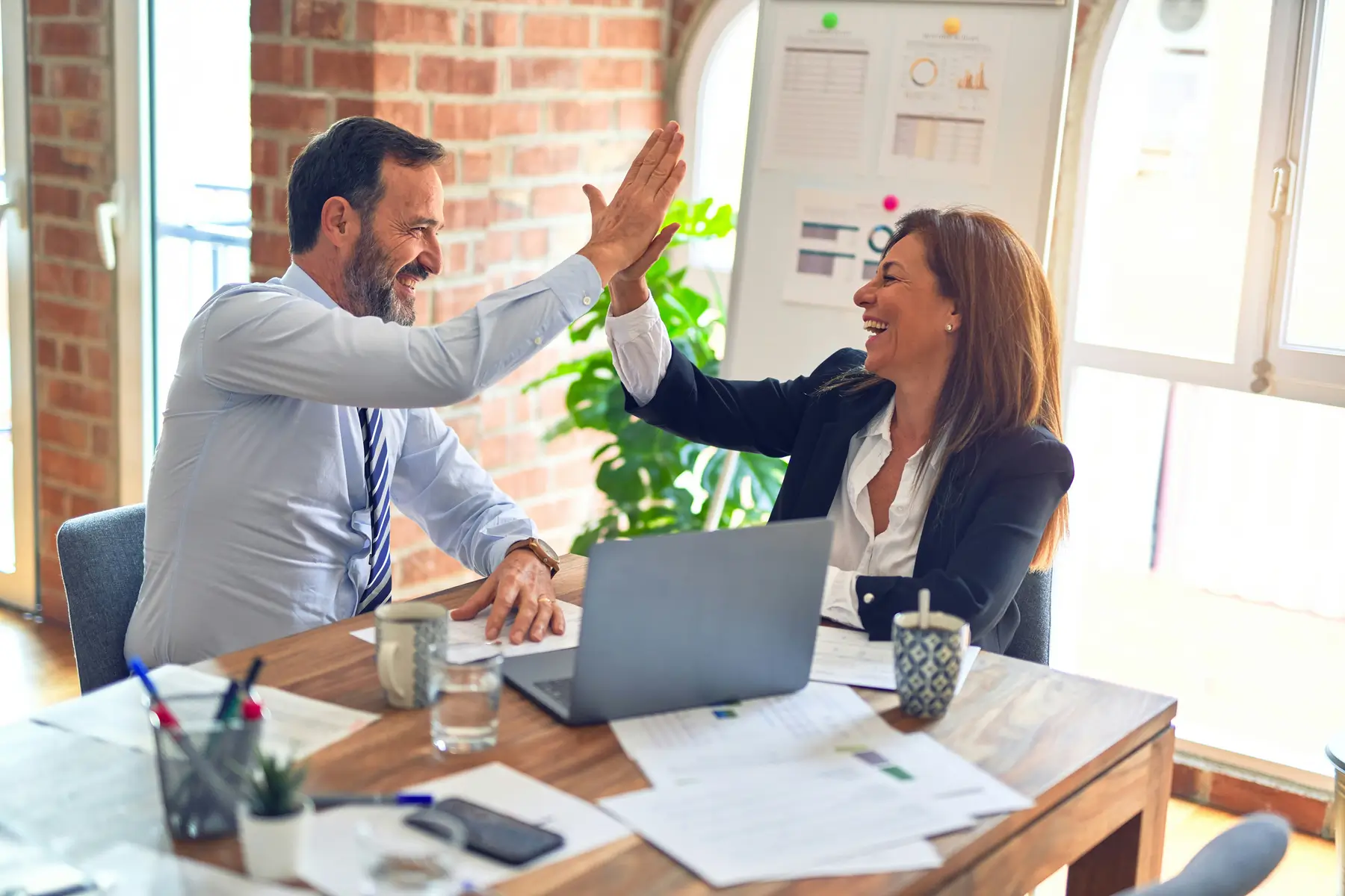 Two colleagues sitting at a table in an office high-five each other, smiling. A laptop, papers, and coffee mugs hint at their shared dedication to bookkeeping. In the background, a plant and charts create a lively atmosphere that underscores their commitment to excellent services.