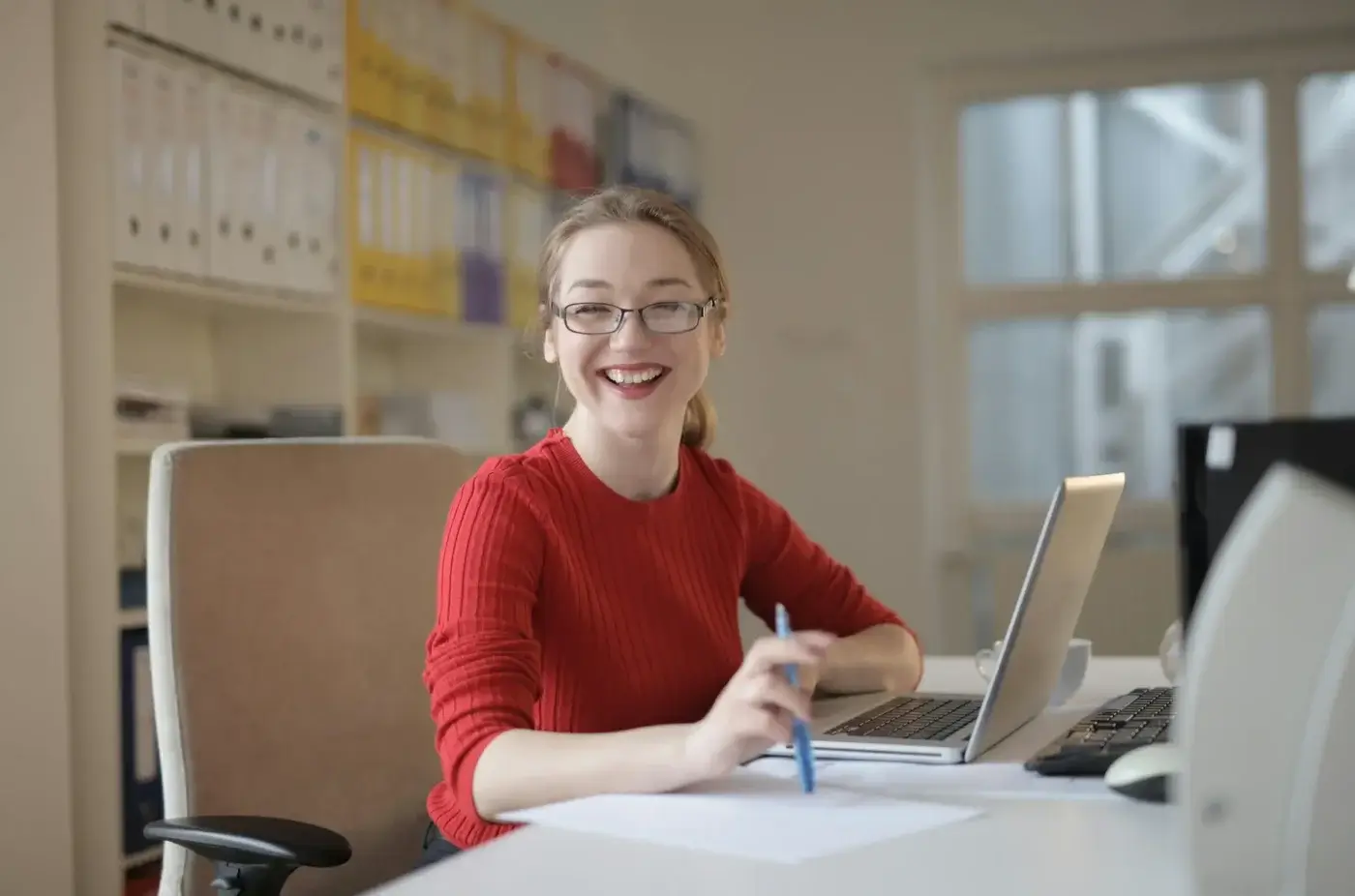 A smiling small business owner in glasses and a red sweater sits at a desk with a laptop and papers, holding a pen, ready to tackle bookkeeping tasks in a bright office environment.