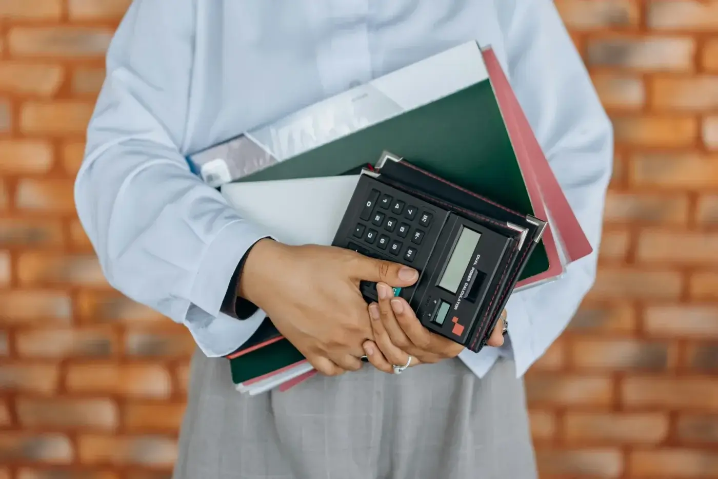 A person holding folders, documents, and a calculator against a brick wall background, representing bookkeeping for service-based businesses.