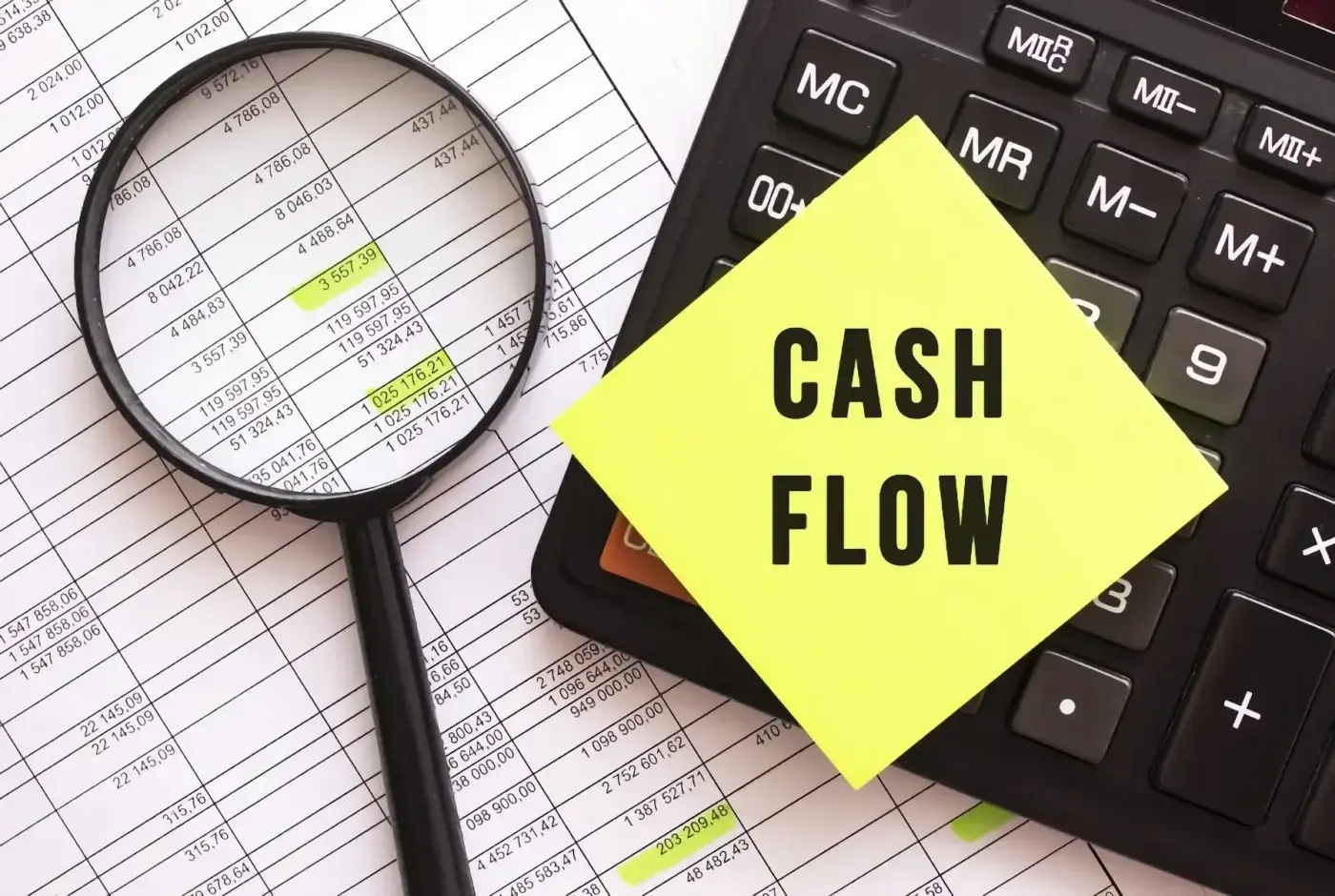 A calculator and magnifying glass rest on financial spreadsheets, with a yellow sticky note labeled "CASH FLOW" in bold letters atop the calculator, highlighting the importance of tracking business cash flow. Some spreadsheet numbers are highlighted.