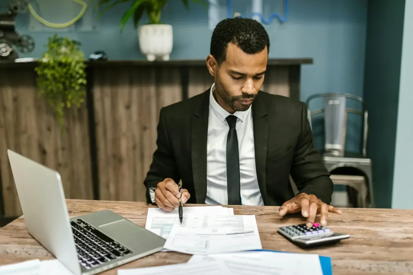 A man in a black suit sits at a desk with papers, a laptop, and a calculator, focusing on calculations in an office setting with a wooden counter and plants in the background.