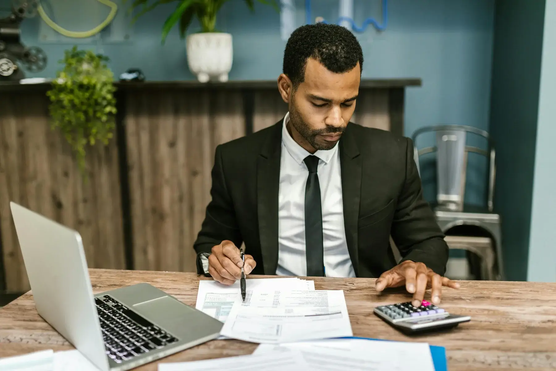 A man in a black suit sits at a desk with papers, a laptop, and a calculator, focusing on calculations in an office setting with a wooden counter and plants in the background.