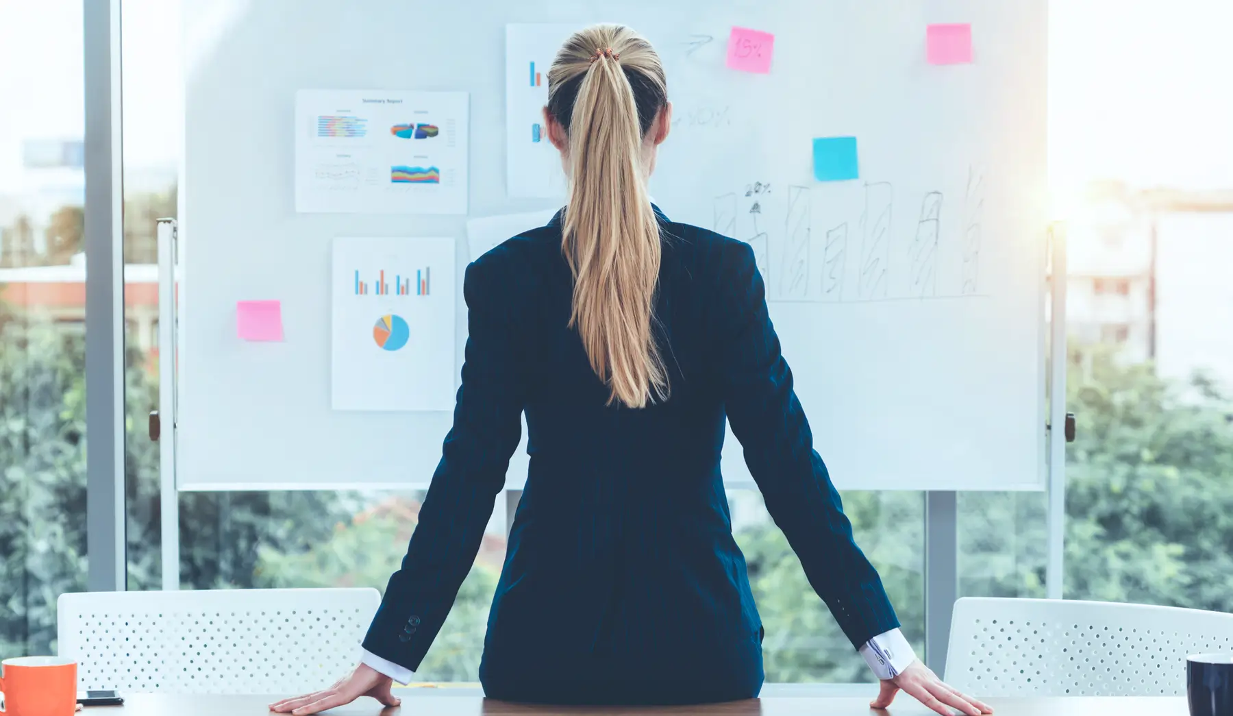 A person in a suit with long hair stands facing a whiteboard covered in graphs and sticky notes, strategizing online bookkeeping solutions for Canada in a brightly lit office area.