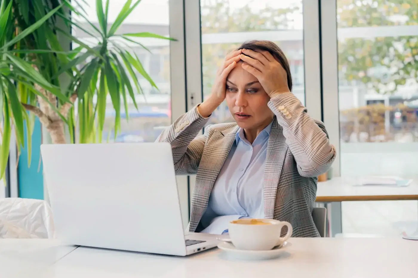 A woman in business attire sits at a table with a laptop, holding her head in distress—perhaps noticing warning signs that her bookkeeping system is costing her money, with a cup of coffee and large windows in the background.