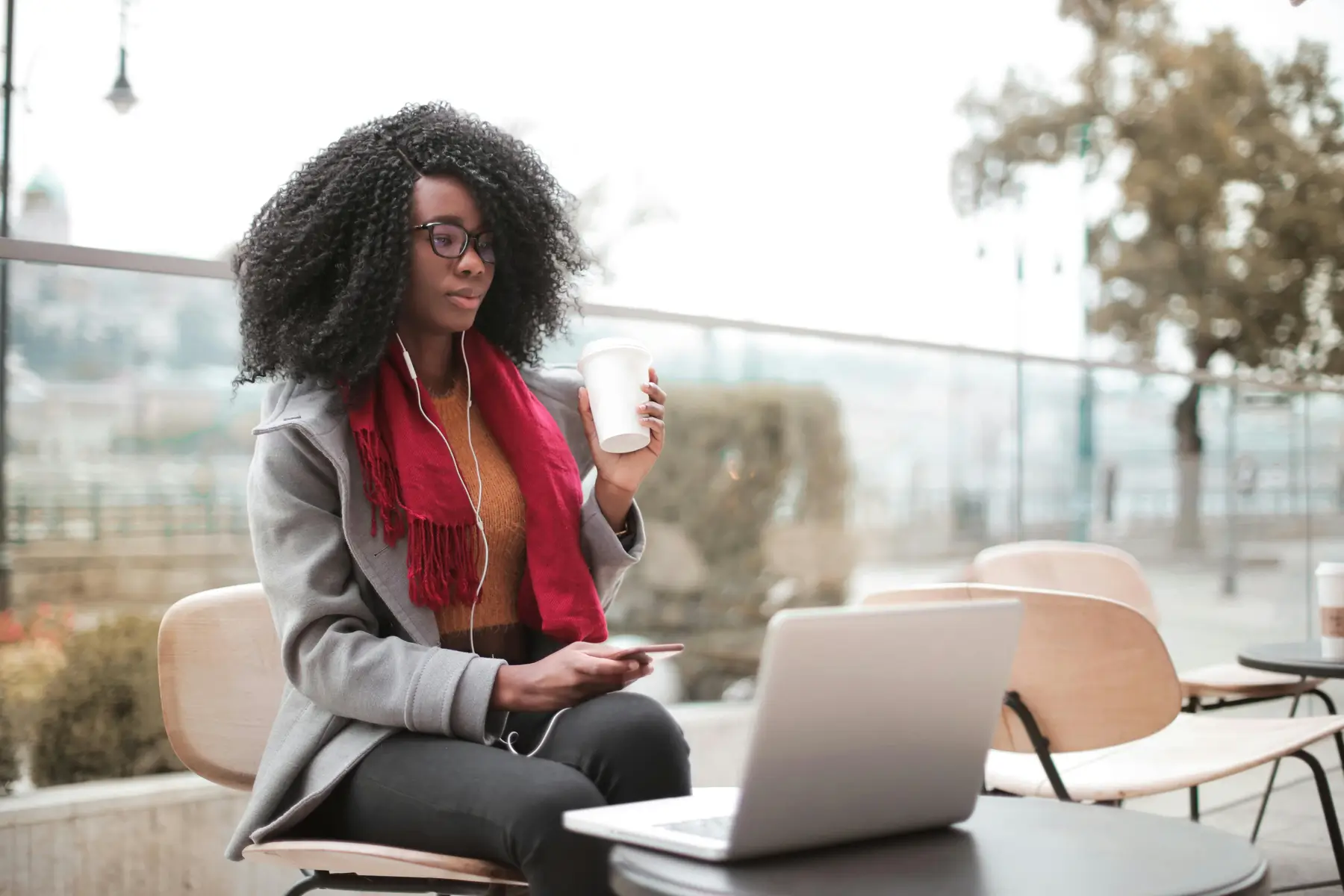 A woman with curly hair and glasses sits outdoors at a café table, holding a coffee cup and using her laptop. Earphones in place, she listens intently—perhaps to tips on how freelancers can stay organized. A red scarf adds color to her gray coat.