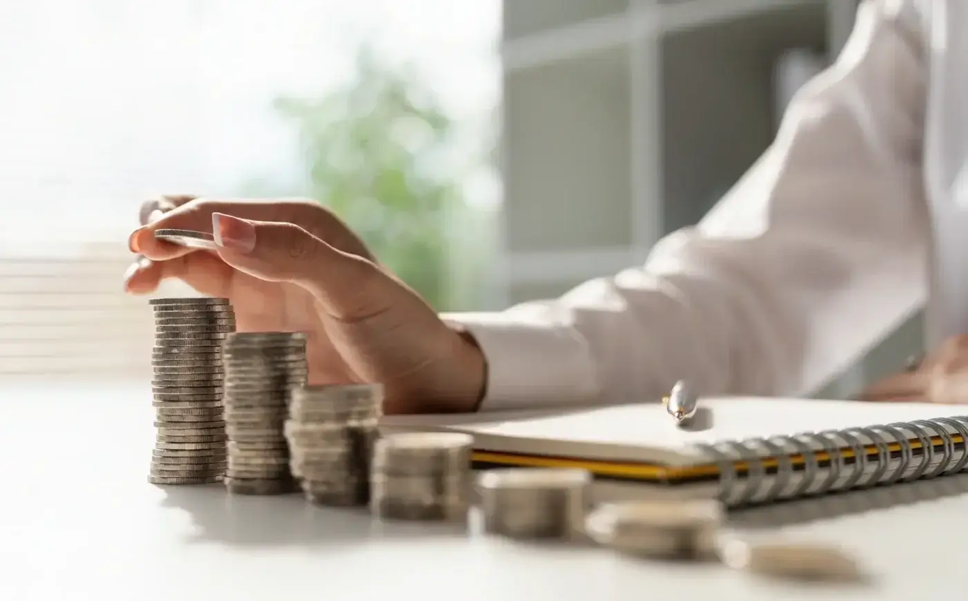 A person stacks coins into tall piles on a desk, with a notebook and pen nearby, symbolizing saving money or financial planning. Sunlight and blurred indoor background complete the scene.
