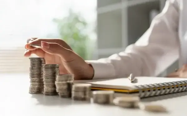 A person stacks coins into tall piles on a desk, with a notebook and pen nearby, symbolizing saving money or financial planning. Sunlight and blurred indoor background complete the scene.
