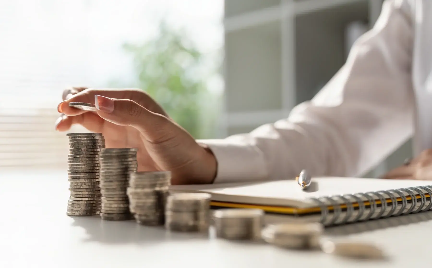 A person stacks coins into tall piles on a desk, with a notebook and pen nearby, symbolizing saving money or financial planning. Sunlight and blurred indoor background complete the scene.