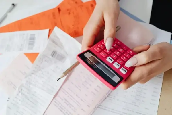 Hands holding a pink calculator above a table scattered with receipts, bills, a pencil, and orange documents, suggesting financial calculations or budgeting.