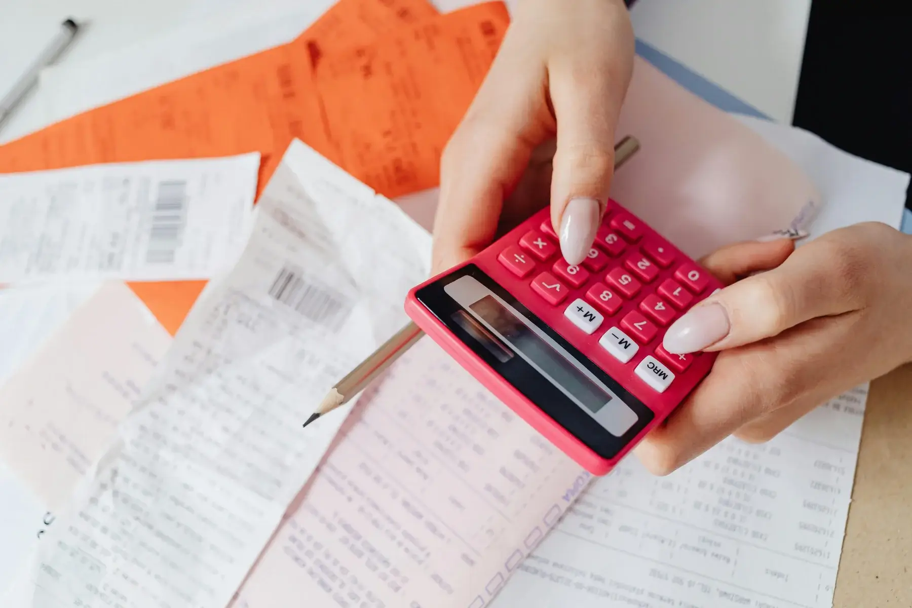 Hands holding a pink calculator above a table scattered with receipts, bills, a pencil, and orange documents, suggesting financial calculations or budgeting.