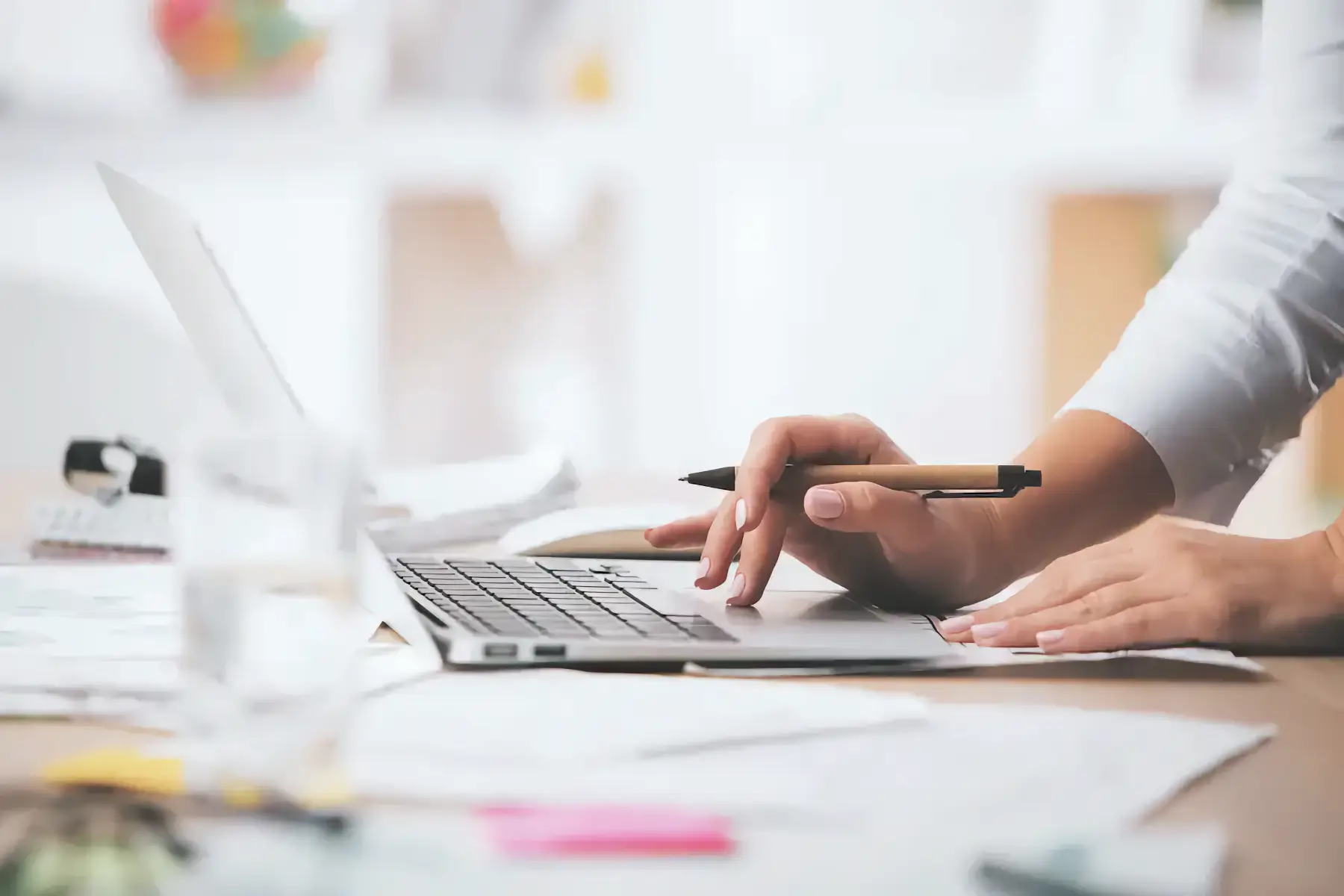 Close-up of a person using a laptop at a cluttered desk, holding a pen with papers, a glass, and a blurred bookshelf in the background.