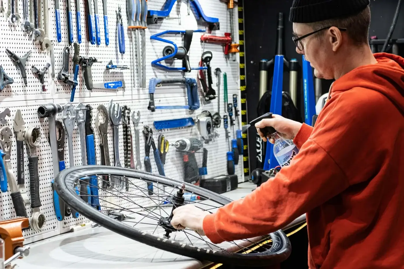Person in an orange hoodie cleans a bicycle wheel with spray in a workshop, with various tools hanging on a pegboard—perfect for service-based business owners tracking business expenses Canada.