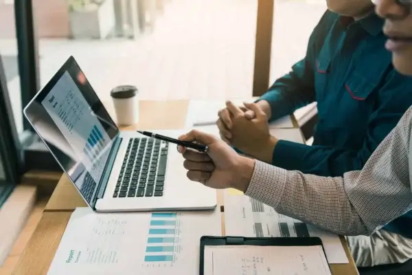 Two people at a wooden table review charts on a laptop screen, with printed charts and a notepad nearby. One person points at the screen with a pen, exploring virtual bookkeeping services in Canada for more efficient financial management.