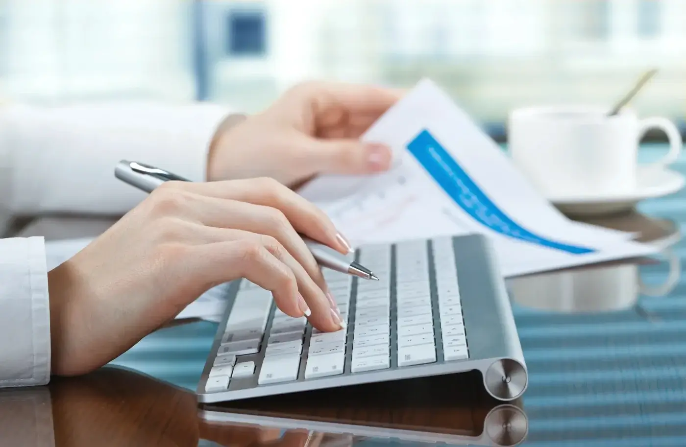 A Bookkeeper types on a wireless keyboard while holding papers in one hand. A pen rests between their fingers, and a white cup and saucer sit on the desk next to them—capturing the essence of what bookkeeping involves daily.